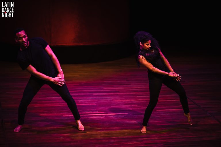 Twee dansers in zwarte kleding voeren een moderne dans uit op een levendig verlichte achtergrond.