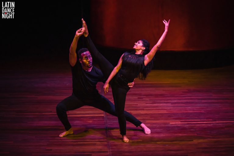Een duo dansers voert een expressieve pose uit op een houten vloer, met een kleurrijke achtergrond.