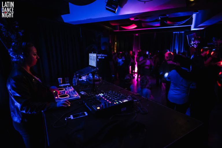 Een avondclubomgeving met een DJ-set op de voorgrond en mensen die dansen in een sfeervolle, blauw-paarse verlichting.