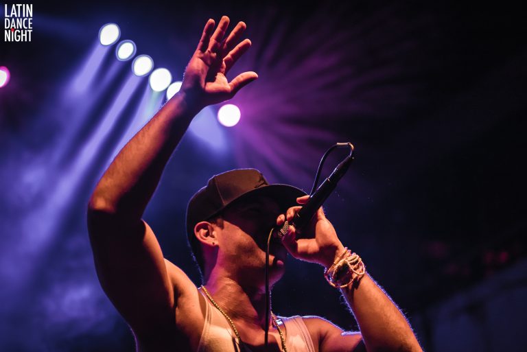 Een performer houdt een microfoon vast en steekt zijn hand omhoog tijdens een live optreden met kleurrijke verlichting op de achtergrond.