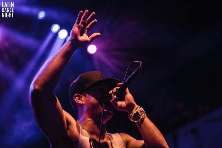 Een artiest die op het podium zingt met een microfoon in de hand, terwijl hij zijn andere hand omhoog steekt.