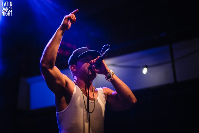 Een performer op het podium houdt een microfoon vast en steekt zijn arm naar boven, omgeven door blauwe lichten.