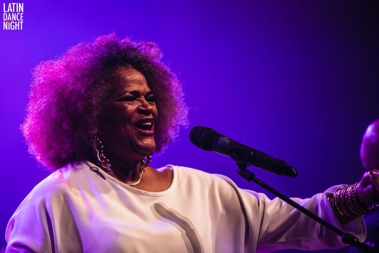 Vrouw met krullend haar zingt enthousiast in een paarse belichting, terwijl ze een microfoon vasthoudt.