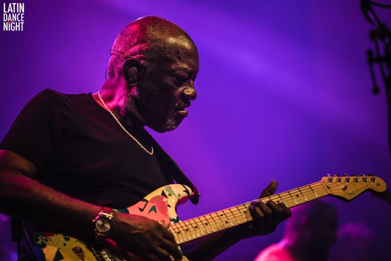Een muzikant speelt gitaar met intense expressie op een podium verlicht door paarse lichten.