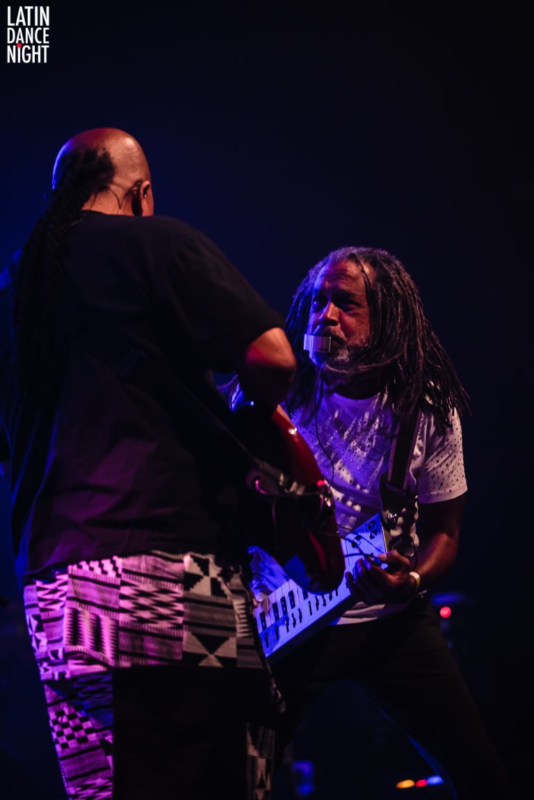 Twee muzikanten spelen samen op het podium, waarbij één de gitaar bespeelt en de ander op hem gericht is.