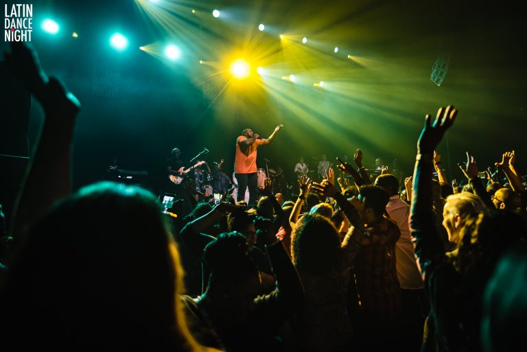 Een levendig concert met een performer op het podium en een enthousiast publiek dat zijn handen omhoog steekt onder kleurrijke lichten.