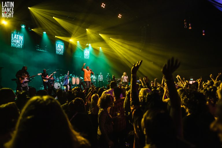 Een groot publiek met opgestoken handen geniet van een live concert onder kleurrijke lichten.