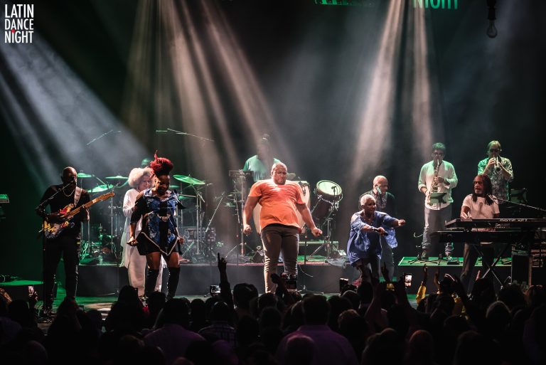 Zes performers staan op een podium met kleurrijke verlichting tijdens een live optreden.