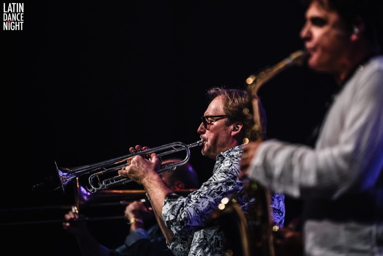 Twee muzikanten spelen krachtige blaasinstrumenten op een podium met een donkere achtergrond.