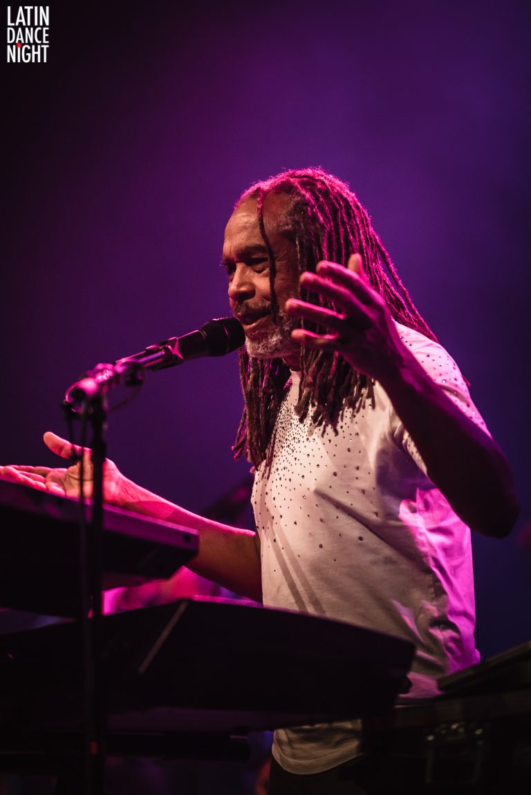 Een muzikant zingt en speelt op een keyboard, met kleurrijke belichting op de achtergrond.
