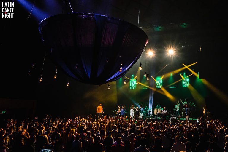 Een drukke concertzaal met een publiek dat naar een liveoptreden kijkt, verlicht door kleurrijke lichten.