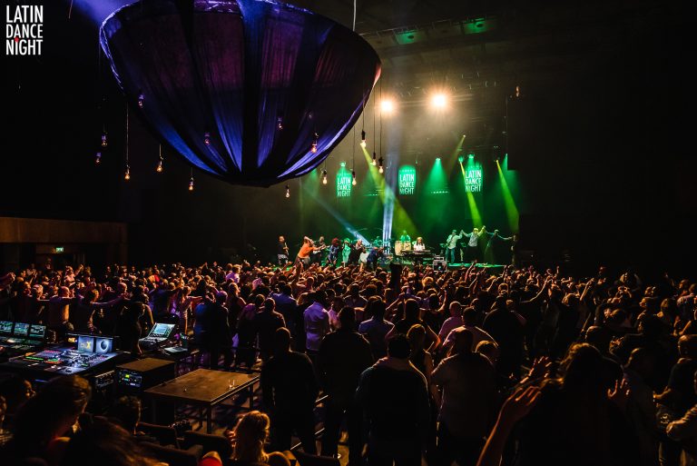 Een drukke concertzaal met een groot aantal mensen die naar een live band kijken, omringd door kleurrijke verlichting.
