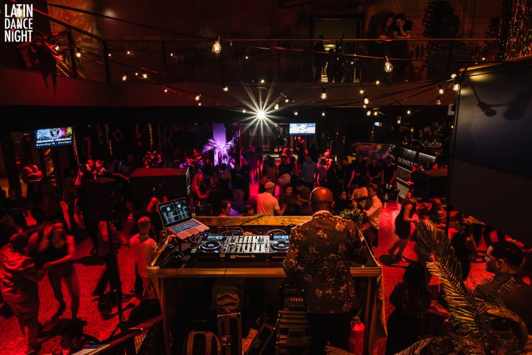 Een drukke dansvloer met een DJ-set op de voorgrond en kleurrijke lichten die de sfeer van het feest benadrukken.