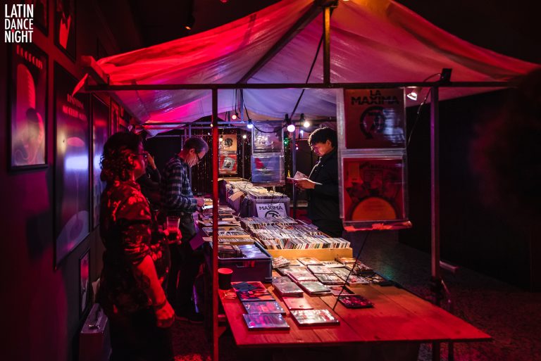 Een drukke kraam op een markt met kleurrijke verlichting en bezoekers die naar de beschikbare items kijken.