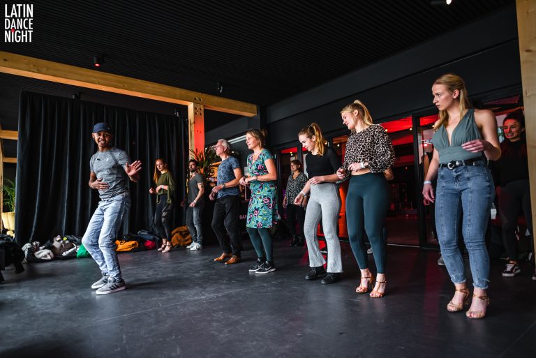 Een groep mensen volgt een dansles in een studio met een zwarte vloer en een houten achtergrond.