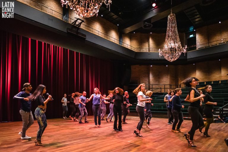 Een groep mensen danst in een stijlvolle zaal met een houten vloer en kroonluchters.