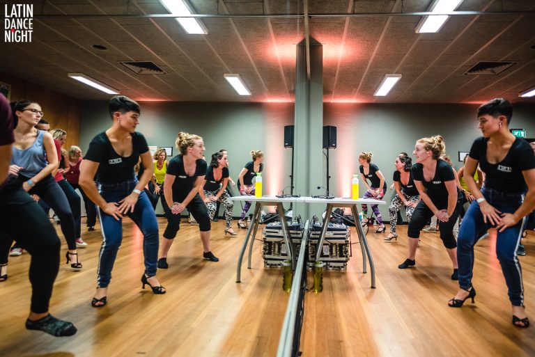 Groep mensen in zwarte shirts die samen dansen in een studio met spiegels.
