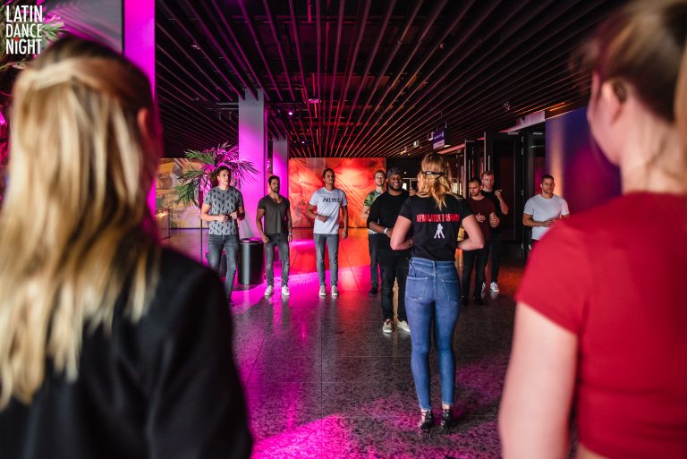 Een groep mensen staat in een modern, verlicht interieur met kleurrijke verlichting en luistert naar een spreker.