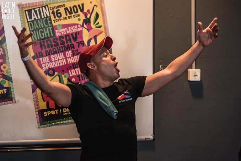 Een persoon met een rode pet en een zwart shirt staat enthousiast met uitgestrekte armen, terwijl hij zingt of iets voordraagt voor een kleurrijk poster.