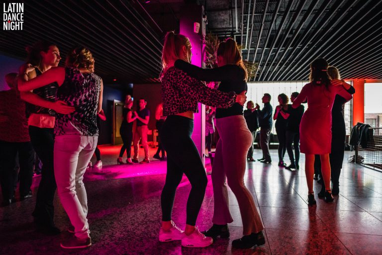 Twee vrouwen dansen samen onder sfeervolle lichten in een drukke dansomgeving.