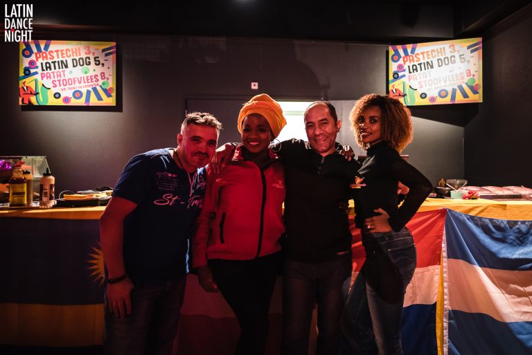 Vier mensen poseren samen voor een kleurrijke achtergrond met vlaggen.