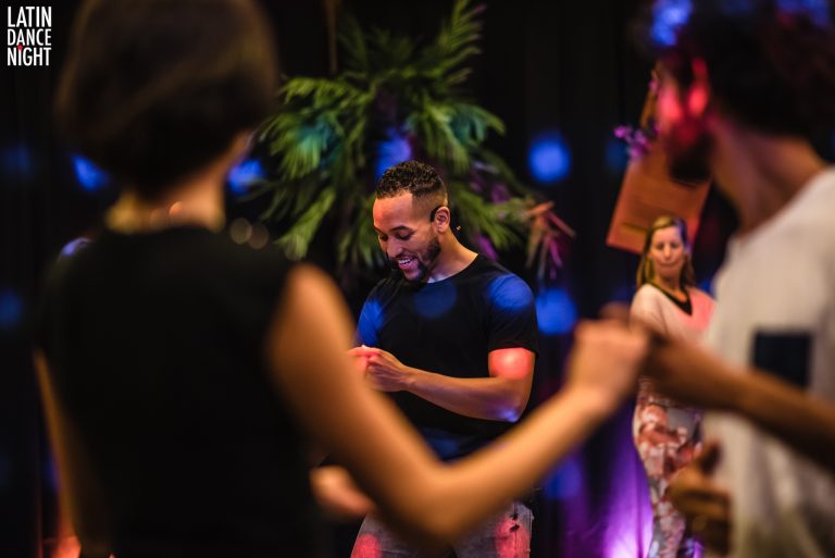 Een groep mensen danst op een feest, omringd door kleurrijke verlichting en groene planten.