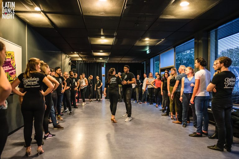 Een dansgroep oefent in een grote zaal met rijen mensen die naar een danspaar kijken.