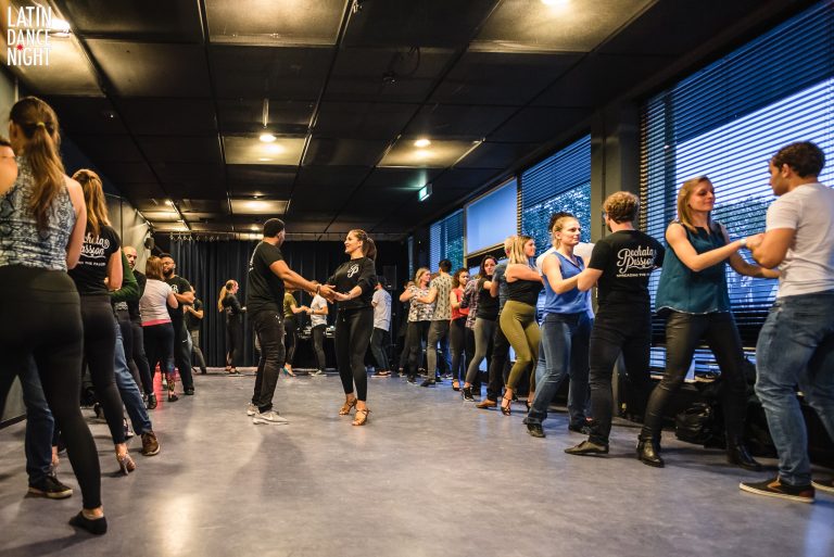 Een groep mensen danst in paren in een moderne dansstudio met grote ramen.