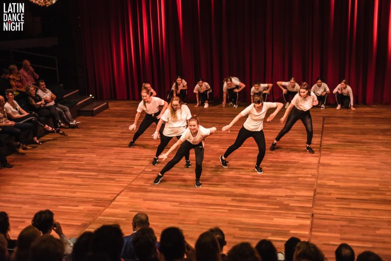 Een groep dansers voert een choreografie uit op een podium met houten vloeren en een achterwand van rode gordijnen.