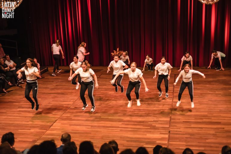 Een groep dansers voert een energieke dansroutine uit op een podium met een houten vloer en een rode achtergrond.