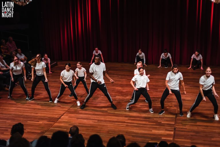 Een groep dansers in witte t-shirts en zwarte broeken voert een choreografie uit op een houten podium.