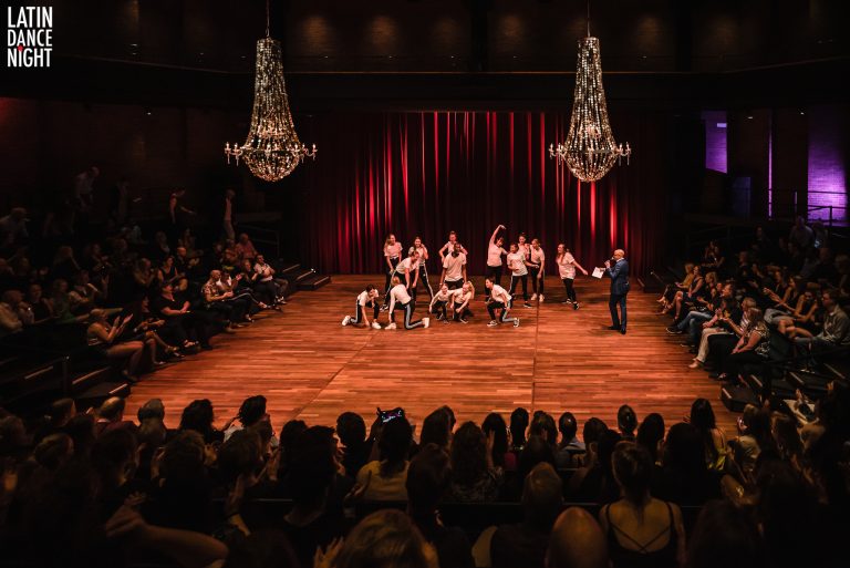 Een dansvoorstelling met performers op een podium, omringd door een publiek onder chandeliers.