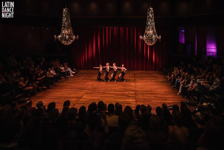 Een dansvoorstelling op een sfeervol verlichte vloer met een publiek in de achtergrond.