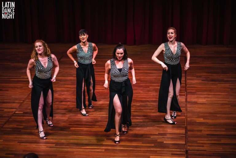 Vijf vrouwelijke dansers in glitterende kostuums voeren een choreografie uit op een podium.