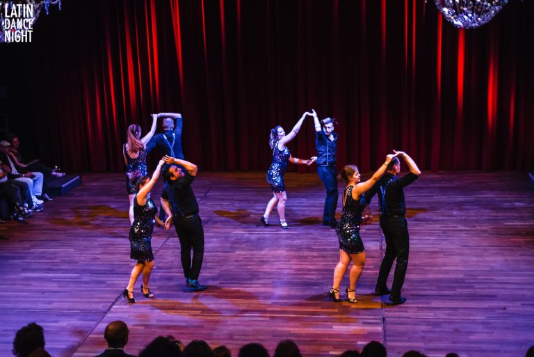 Zes dansers in zwarte outfits voeren een choreografie uit op een sfeervol verlichte dansvloer.