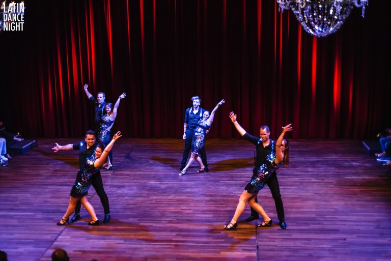 Een groep dansers in stijlvolle outfits performt op een podium met een rode achtergrond en een discobal boven hen.