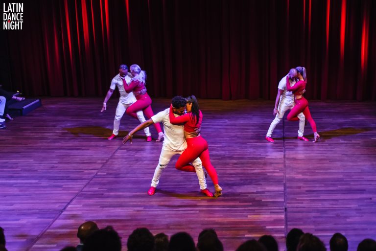 Een dansgroep voert een energieke choreografie uit op een podium met houten vloer en rode gordijnen op de achtergrond.
