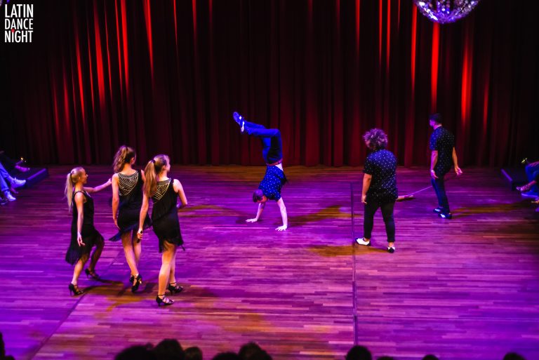 Groep dansers voert een dynamische routine uit op een podium met een kleurrijke verlichting.