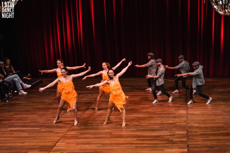 Groep dansers in oranje kostuums presenteert een energieke choreografie op een podium met een houten vloer en rode achtergrond.