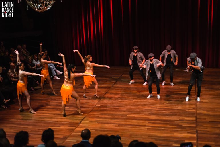 Groep dansers in levendige kostuums optredend op een houten podium met een rode achtergrond.