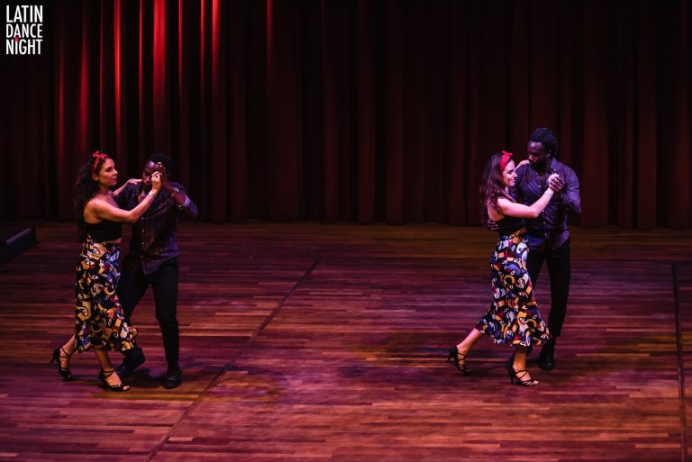 Twee paren dansen samen op een podium met gedempt licht en een warmgekleurde achtergrond.