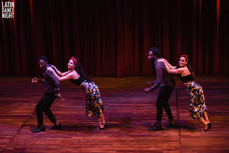 Vijf dansers voeren een energieke choreografie uit op een podium met een houten vloer en een roodachtig achtergronddoek.