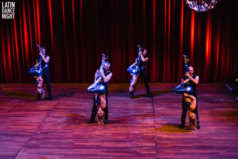 Meerdere dansers in zwarte outfits voeren een choreografie uit op een podium met een rode achtergrond.