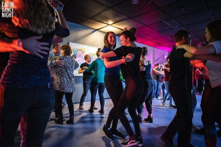 Twee mensen dansen samen in een drukke zaal met kleurrijke verlichting en andere dansers op de achtergrond.