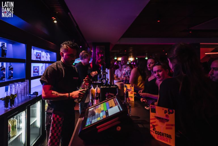 Een drukke bar met barmensen die drankjes bereiden en gasten aan de bar die met elkaar converseren.