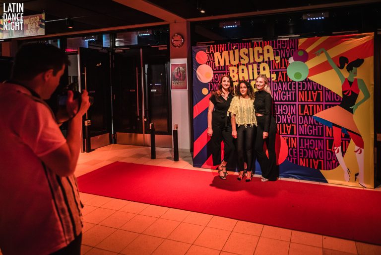 Drie mensen poseren op een rode loper voor een kleurrijke achtergrond met de tekst 