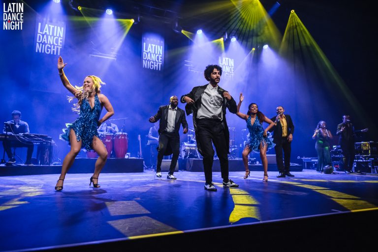 Zes dansers performen op een podium met kleurrijke verlichting tijdens een live show.