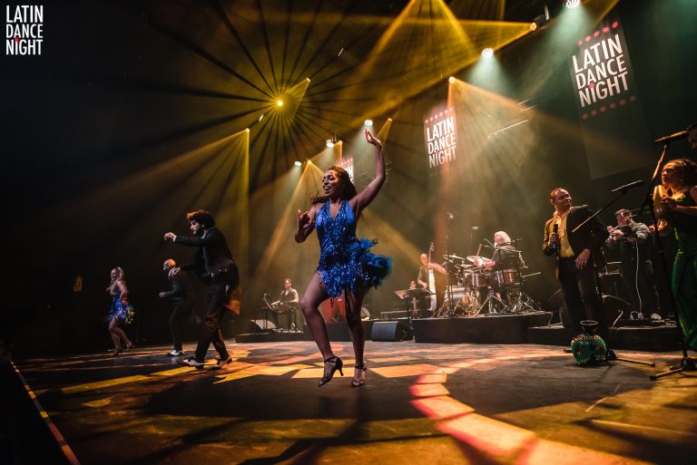 Een levendige uitvoering op het podium met een artiest in een blauwe jurk en kleurrijke lichten.