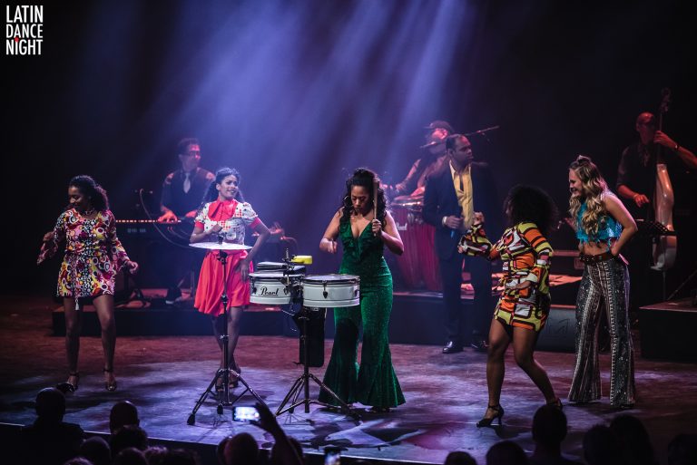 Een live muziekoptreden met verschillende artiesten die instrumenten bespelen en samen muziek maken op het podium.