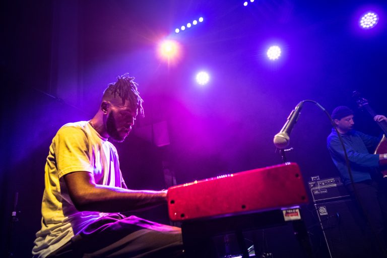 Een muzikant speelt op een rode keyboard tegen een achtergrond van paarse en blauwe lichten.